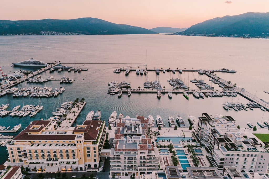 Luxury waterfront hotel with grand Mediterranean-style facade, palm trees and private marina set against Montenegro's mountains at sunset