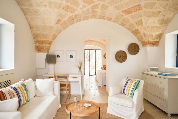 Rustic stone-walled hotel room in Italian countryside, featuring elegant white linens, minimalist decor, and warm natural light through arched window