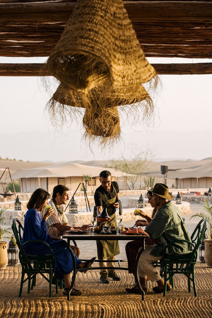 Luxurious desert tent camp with white canvas structures nestled in Moroccan landscape, mountain backdrop and infinity pool at sunset