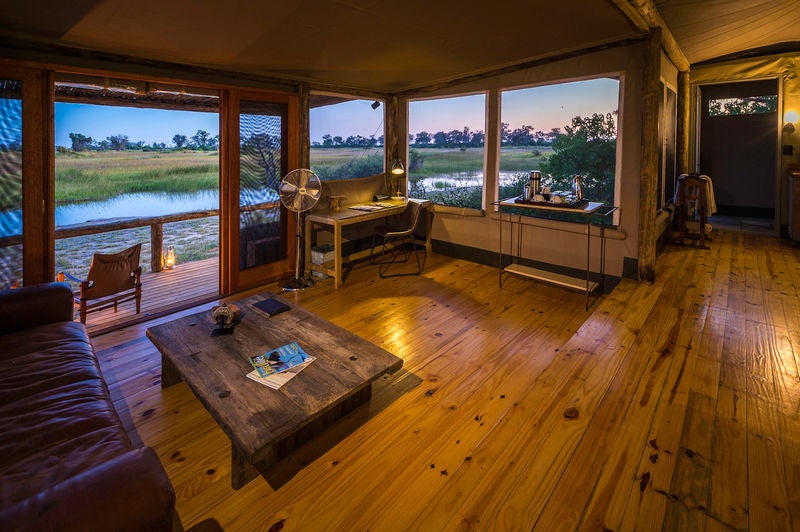 Elevated safari lodge with thatched-roof suites overlooking lush wetlands, featuring private wooden deck and canvas walls at sunset