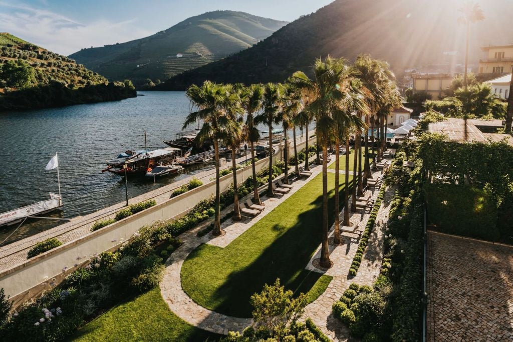 Elegant hillside hotel with stone facade overlooking Douro River valley, surrounded by terraced vineyards and manicured gardens