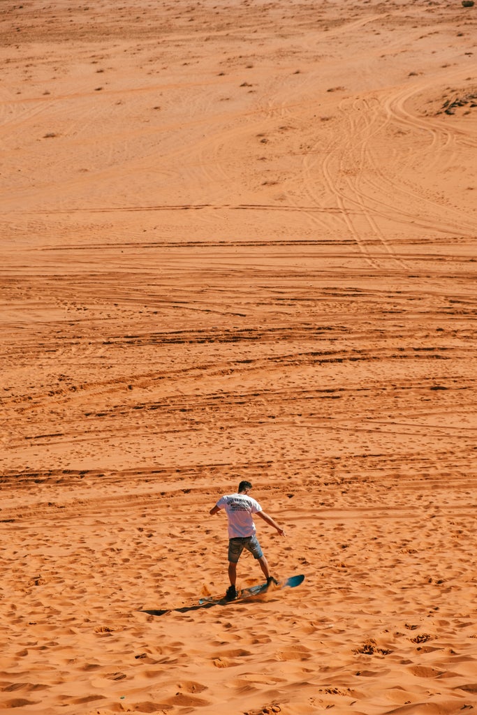 Adventurer glides down golden sand dune in Morocco, wearing premium gear against dramatic desert landscape with distant rocky mountains