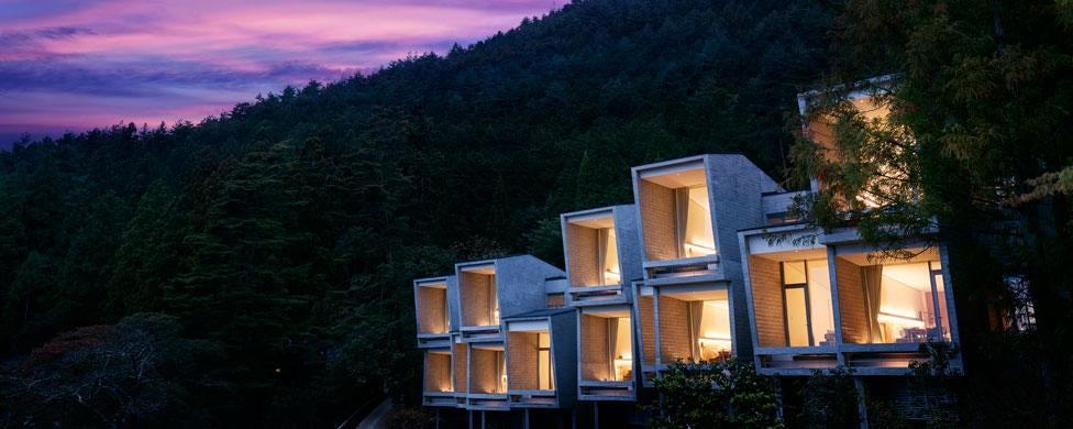 Modern concrete cabin with floor-to-ceiling windows perched on Mount Fuji hillside, offering panoramic views of Lake Kawaguchi and forest