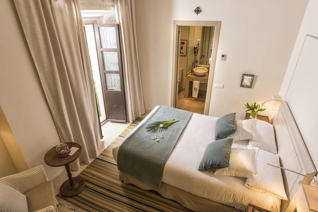 Elegant colonial-style bedroom with ornate wooden furniture, intricate tile floors, and large windows overlooking historic Córdoba's charming architectural landscape