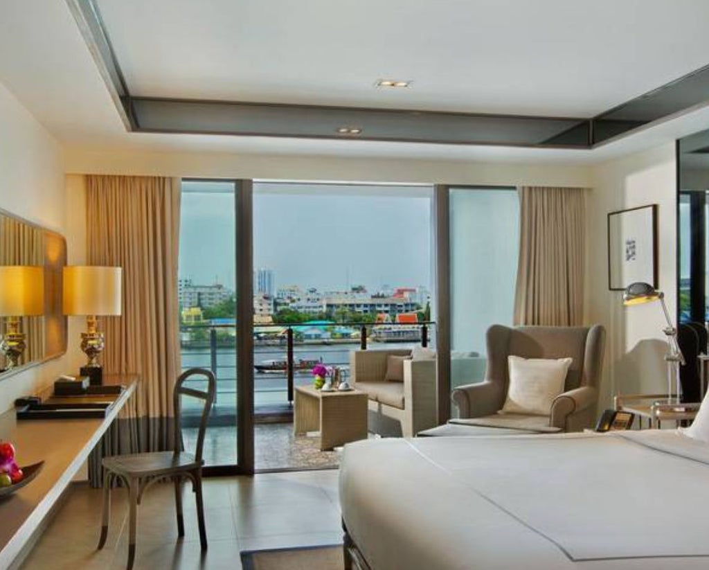 Contemporary Thai hotel room featuring king bed, warm lighting, polished wood floors, white linens, and floor-to-ceiling windows with river views