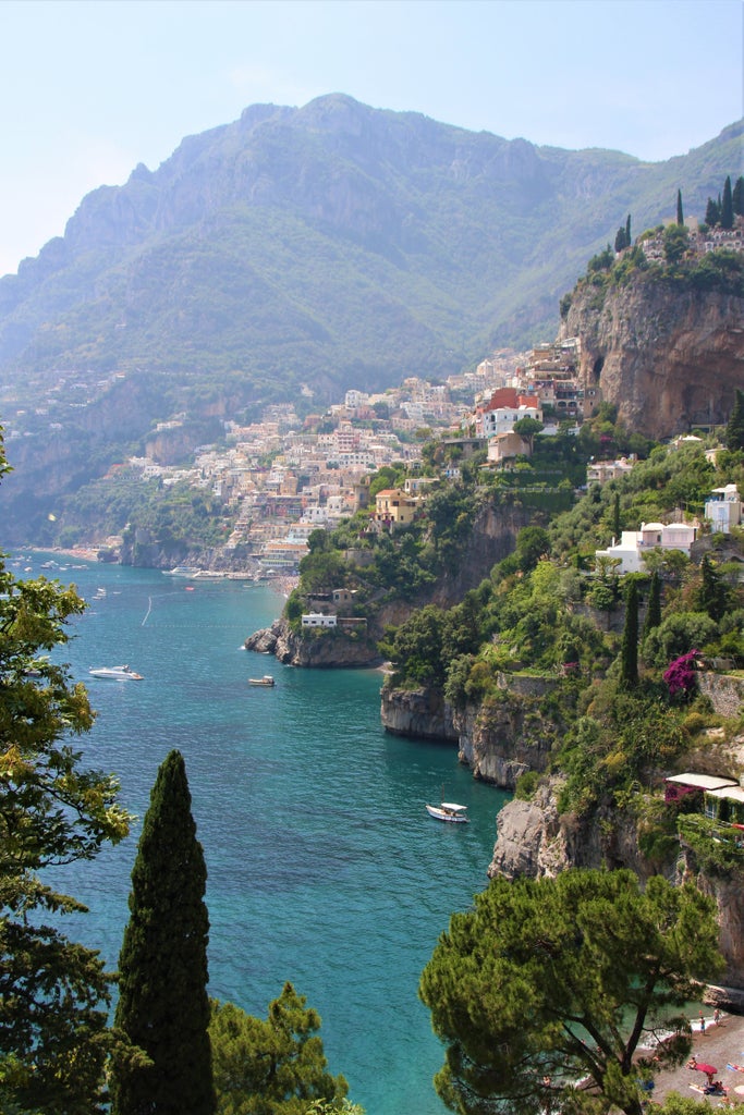 Winding coastal road hugs dramatic cliffs along Mediterranean Sea, with colorful villas and terraced gardens cascading down to turquoise waters