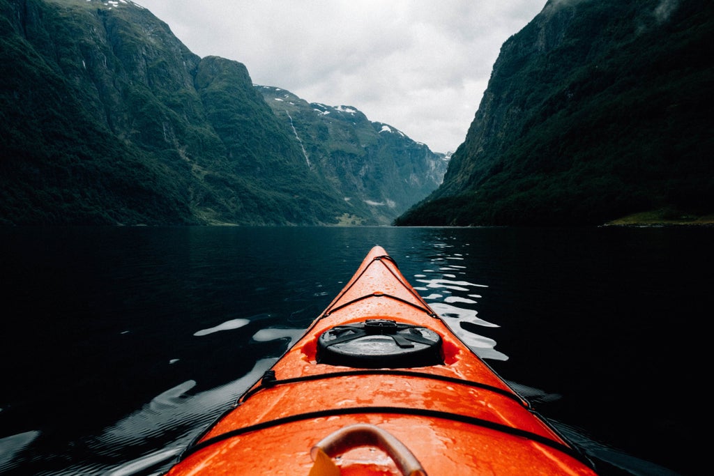 Luxurious sea kayak gliding through serene Norwegian fjord with dramatic steep cliffs, pristine blue waters, and snow-capped mountain peaks in background