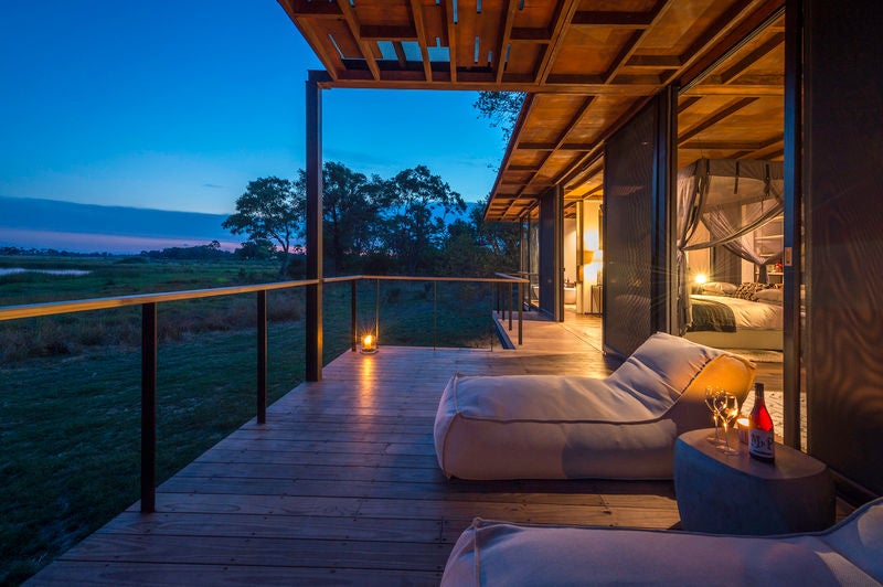 Elevated luxury lodge with thatched roof overlooking pristine Okavango Delta wetlands at sunset, surrounded by acacia trees