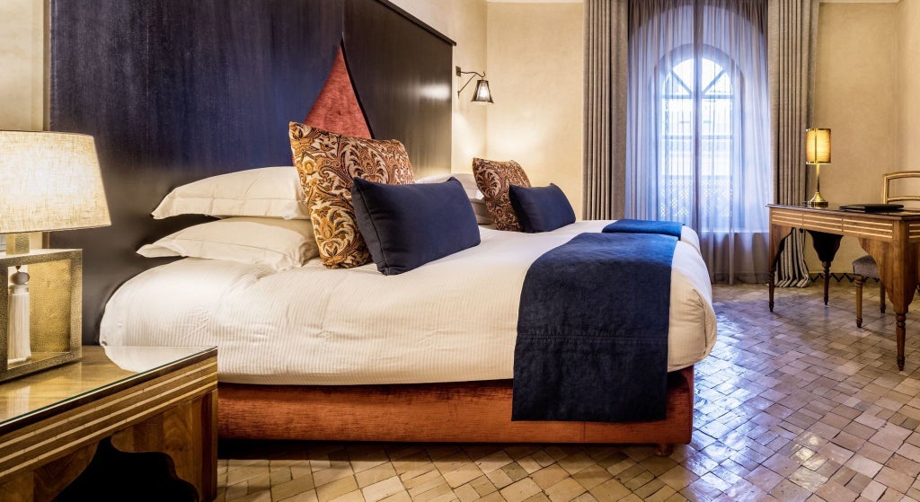 Elegant Moroccan deluxe hotel room with intricate tilework, ornate chandelier, plush bed, and traditional arabesque architectural details in warm earthy tones