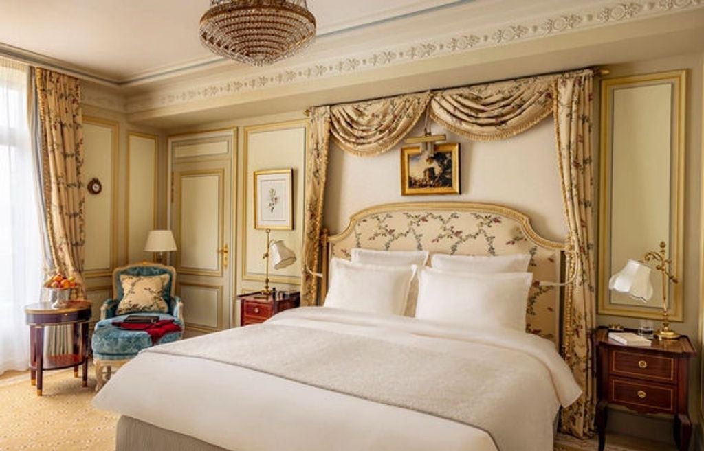 Elegant hotel room with crystal chandelier, ornate gold-framed mirror, Louis XV furniture, and French windows overlooking Paris streets