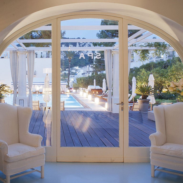 Elegant Mediterranean hotel balcony overlooking Capri's coastline with white columns, manicured gardens and azure sea views at sunset