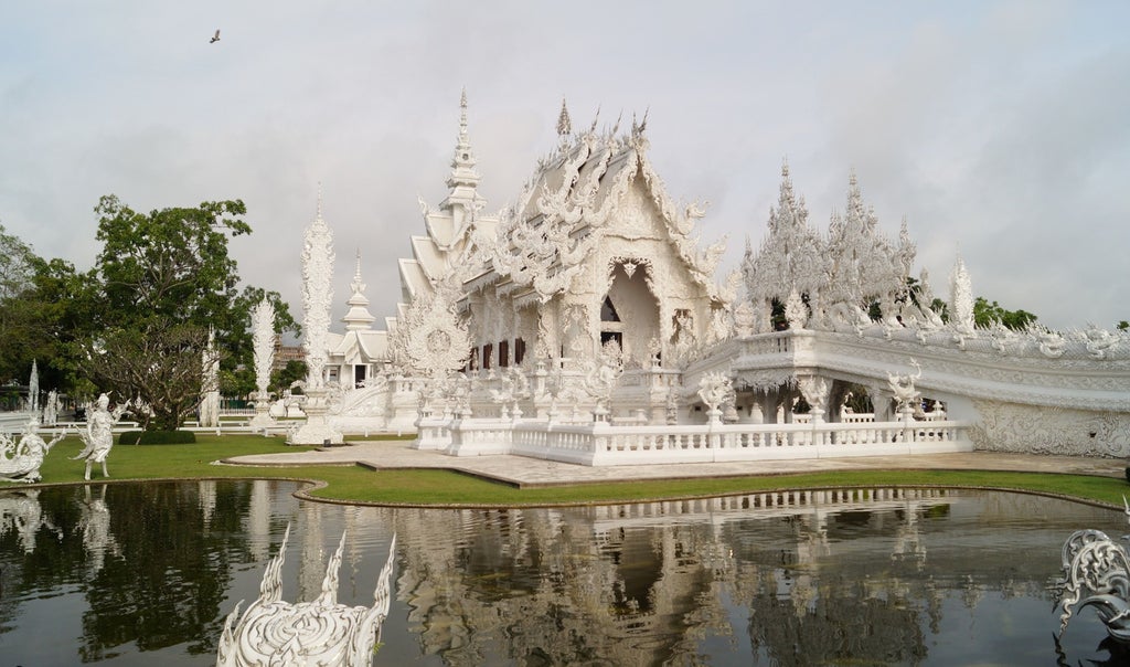 Ornate golden temple spires of Wat Rong Khun rise against lush mountains, reflecting in calm waters during golden hour in Chiang Rai