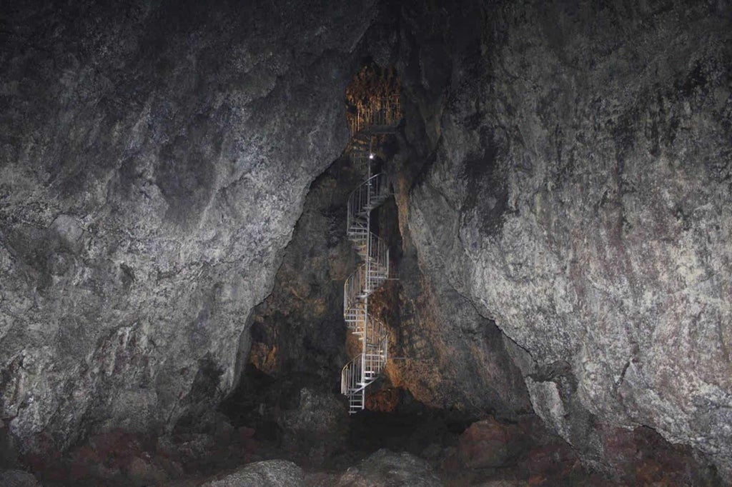 Underground lava cave with stone stairs and railings illuminated by guide's flashlight, revealing dark volcanic rock formations