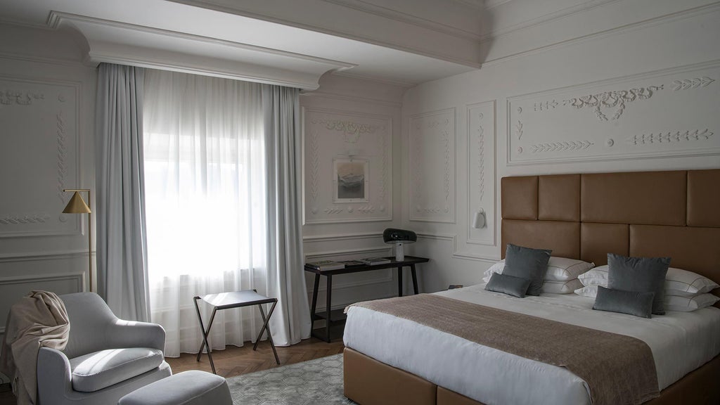 Elegant junior suite in historic Italian hotel, featuring plush white bed, soft neutral tones, antique wooden furnishings, and large arched window with natural light