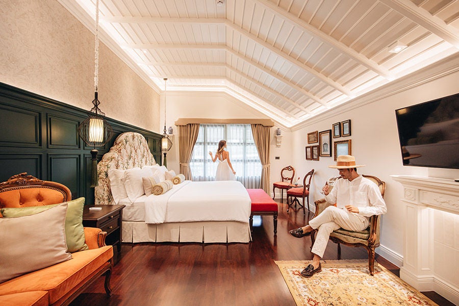 Elegant Lanna-style corner suite with wooden beams, traditional Thai decor, king bed, and private balcony overlooking tropical gardens