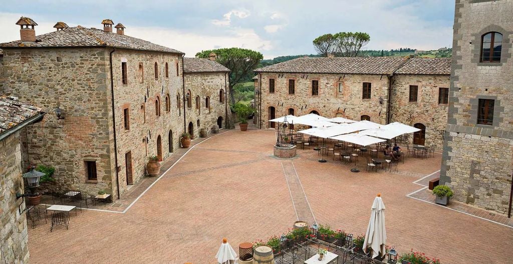 Luxury Tuscan resort with stone buildings surrounding medieval courtyard, terra cotta roofs amid rolling hills at golden hour