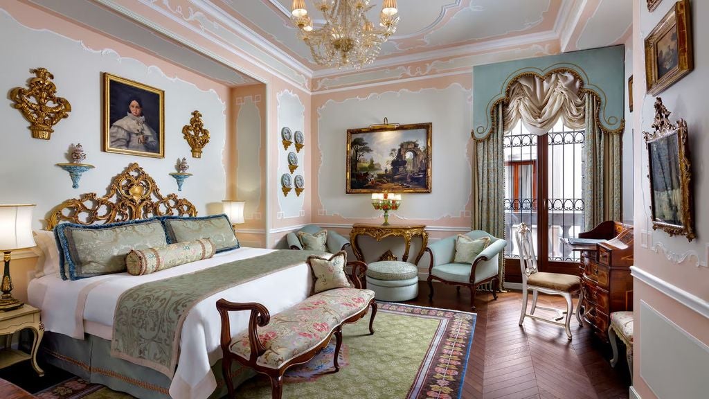 Opulent Venetian suite with rich damask walls, crystal chandelier, antique furnishings, and floor-to-ceiling windows overlooking Grand Canal