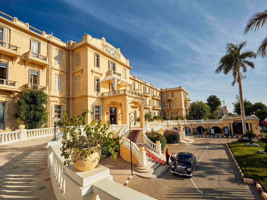 Majestic colonial-style hotel with ornate white façade, grand balconies, and palm trees set against Luxor's bright blue desert sky