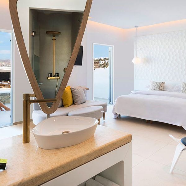 Luxurious white-washed Myconian hotel room with panoramic Aegean Sea view, elegant minimalist design, private balcony overlooking Mykonos coastline at sunrise