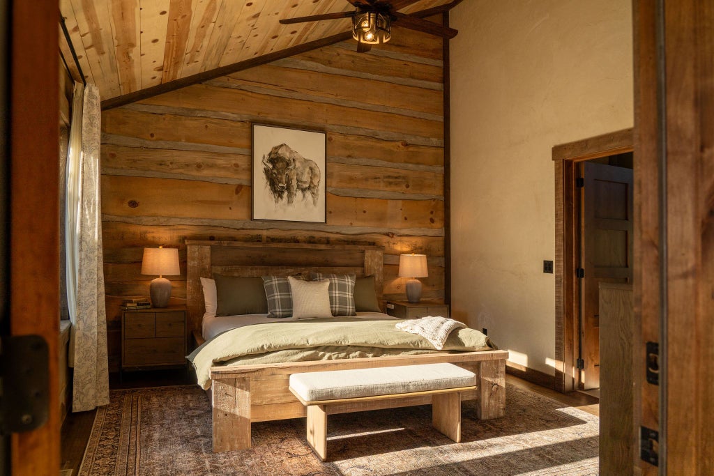 Rustic wooden lodge room with king bed, plush bedding, panoramic mountain view windows, warm earth tones, and elegant southwestern decor at a scenic ranch