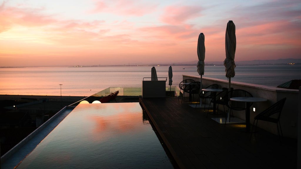 Boutique hotel terrace overlooking Lisbon's historic Alfama district, featuring minimalist design, white furniture, and panoramic cityscape views at sunset