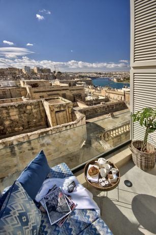 Elegant boutique suite in Malta with soft grey walls, plush white bedding, contemporary minimalist design, and large window overlooking historic scenset architecture