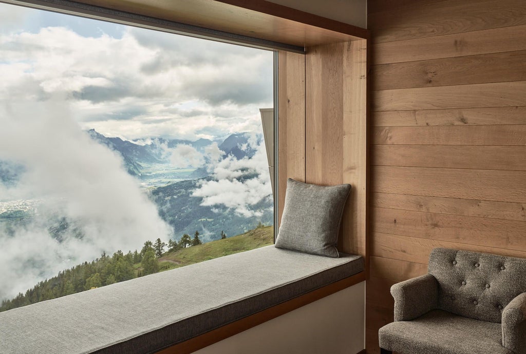 Luxurious alpine hotel room with panoramic mountain views, minimalist wood furnishings, and large windows overlooking Swiss mountain landscape at dusk