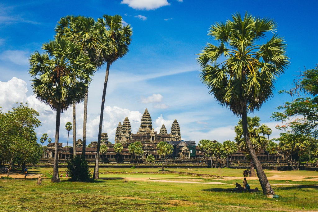 Iconic Angkor Wat towers reflected in still lotus ponds at dawn, with vibrant orange sunrise sky and silhouetted palm trees framing temple