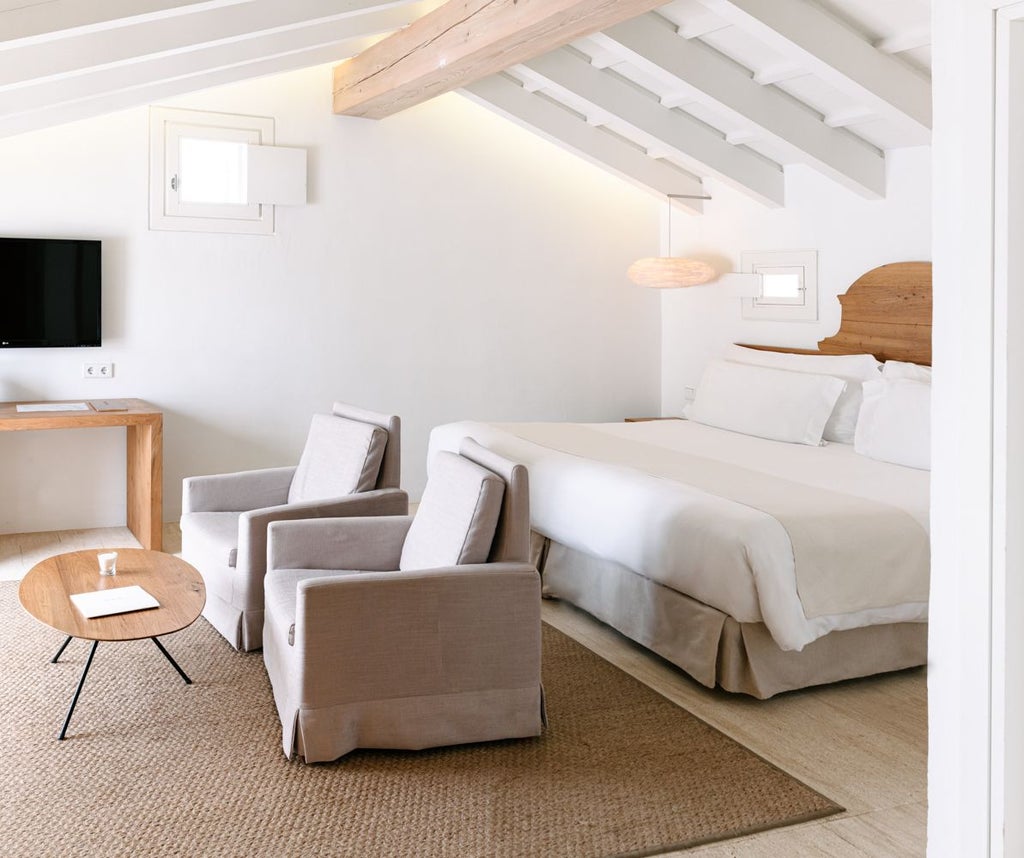 Elegant whitewashed Spanish hotel room with minimalist design, natural light, plush white bedding, and rustic stone wall in Menorca's scenic countryside