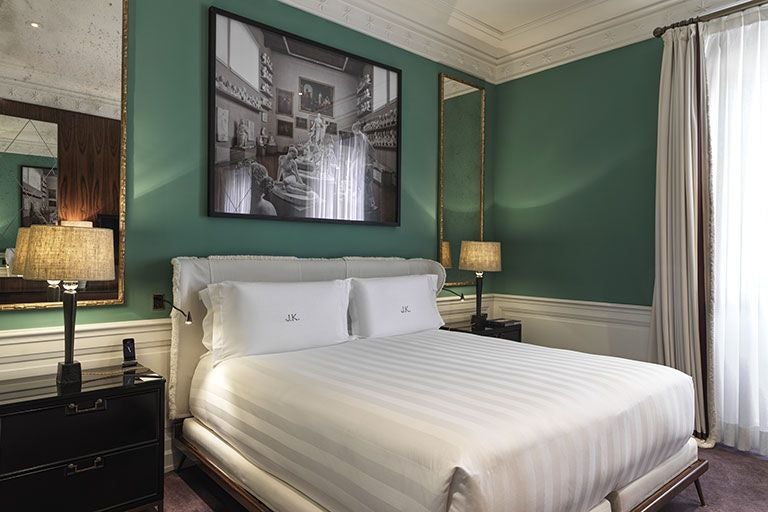 Elegant master bedroom at J.K. Place Roma with plush cream-colored linens, marble accents, and panoramic view of Rome's historic architecture.