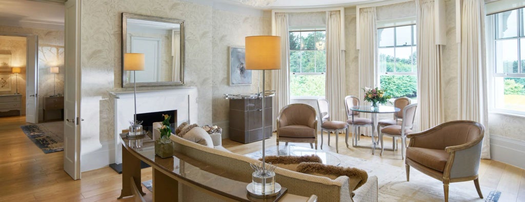 Elegant luxury suite with plush white bedding, ornate chandeliers, classic wood furnishings, and expansive windows overlooking manicured gardens at Coworth Park hotel.