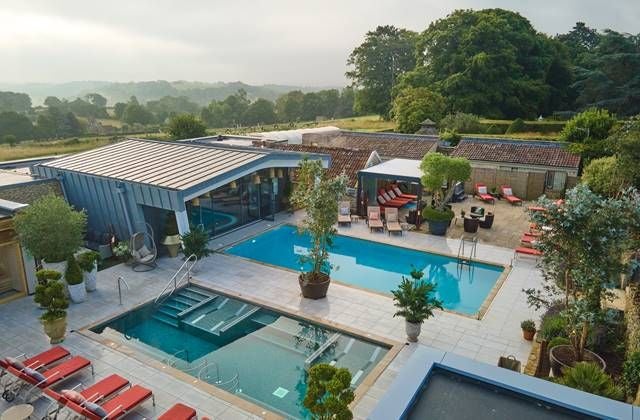 Elegant modern hotel exterior with sleek glass facade, manicured gardens, and luxurious spa wing overlooking scenic UK countryside landscape at dusk