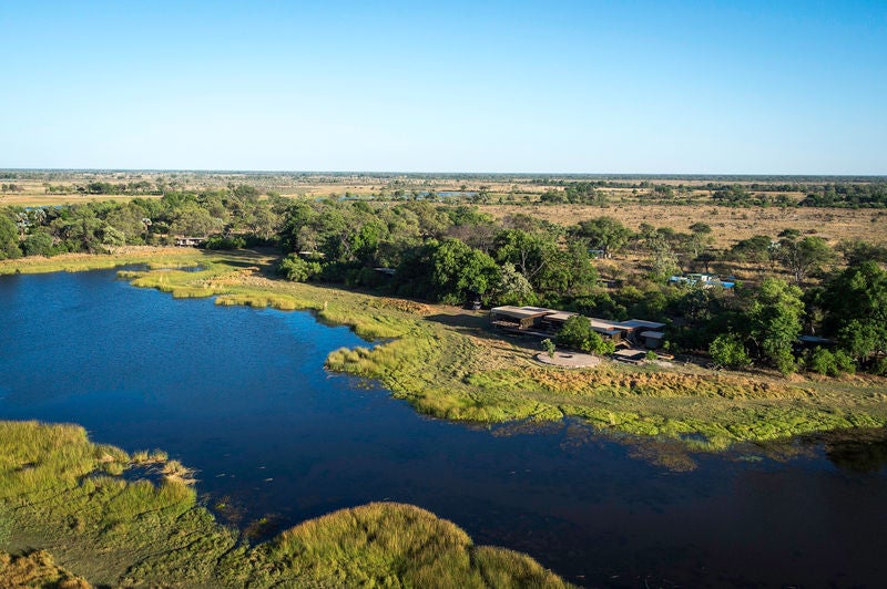 Modern luxury safari lodge with thatched roof overlooking tranquil lagoon, featuring elevated wooden deck and infinity pool