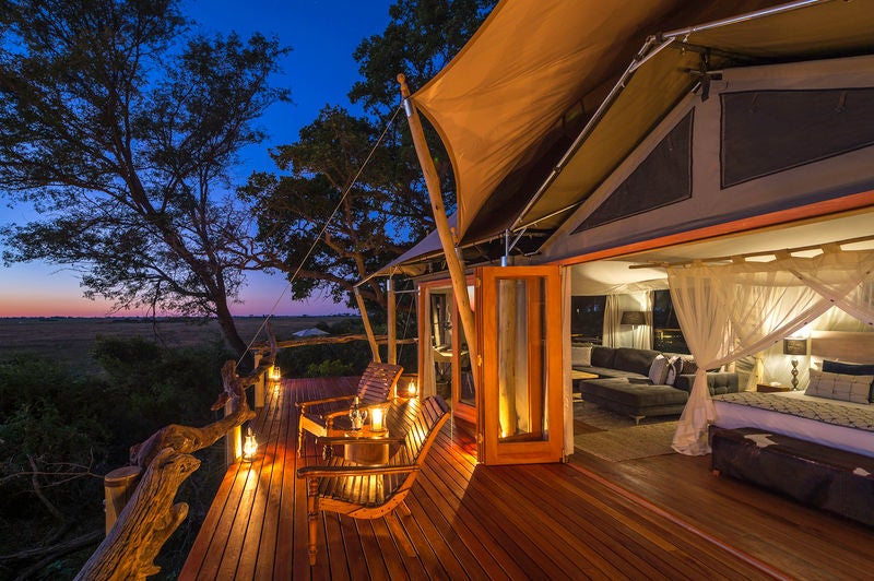 An elevated luxury safari lodge with thatched roof, wooden deck overlooking flooded Okavango Delta plains during sunset