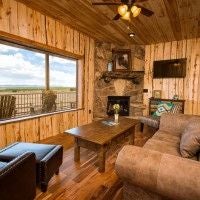 Rustic wooden lodge room with plush king bed, expansive mountain view windows, warm earth-toned decor, and natural stone fireplace at Scenset Mountain Ranch