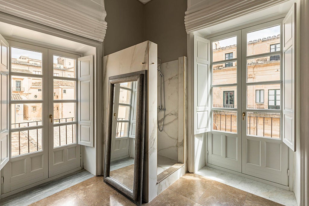 Elegant Sicilian-style deluxe superior hotel room with soft neutral tones, ornate vintage furnishings, and large windows overlooking historic architecture