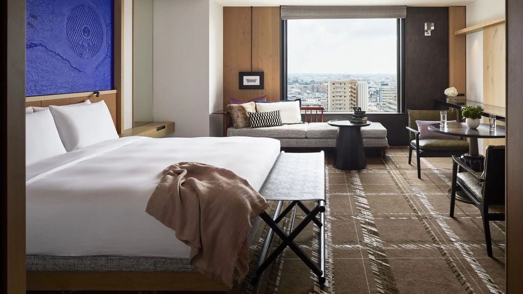 Modern hotel room with plush king bed, floor-to-ceiling windows, sleek wooden furnishings, and panoramic city views in soft natural light