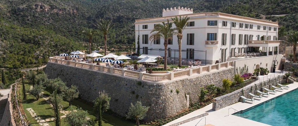 Mediterranean mountainside hotel with stone facades, terraced gardens, and infinity pool overlooking Mallorca's coastline and olive groves