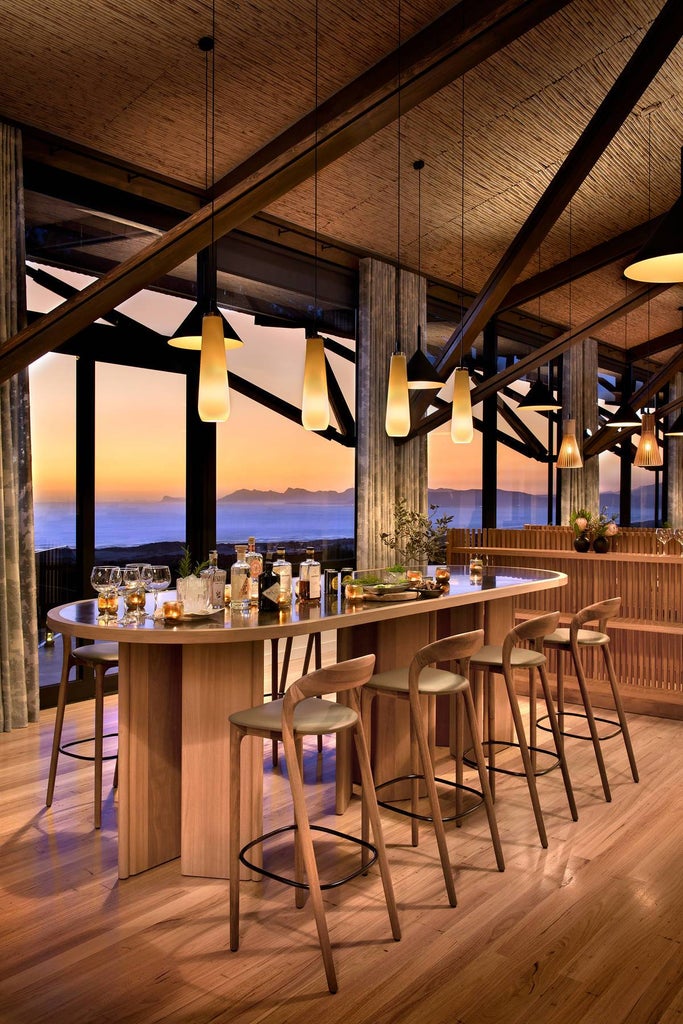 Modern luxury lodge with floor-to-ceiling windows overlooking green hills and ocean, featuring stone walls and wooden deck terrace