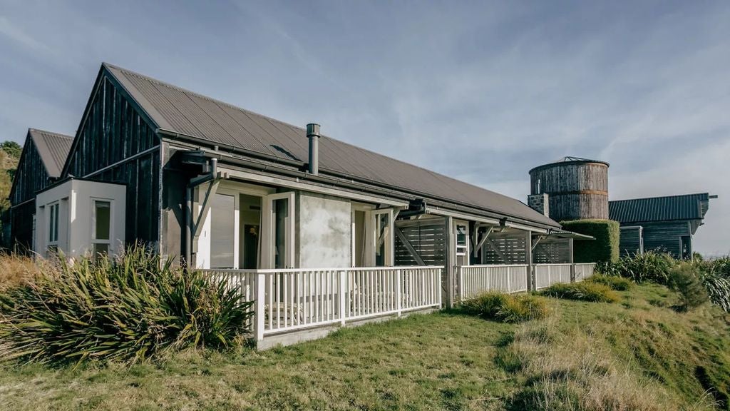 Spacious luxury suite with king bed, rustic wood beams, and floor-to-ceiling windows overlooking New Zealand's dramatic coastline