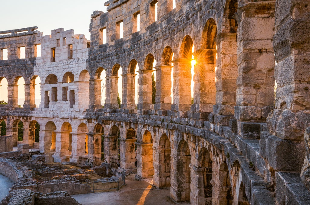 Historic Roman Amphitheater in Pula at sunset with golden light illuminating limestone arches, overlooking turquoise Adriatic waters