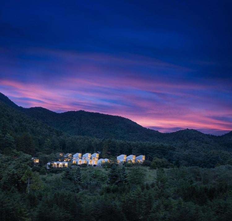 Minimalist luxury cabin with floor-to-ceiling windows overlooking Mount Fuji, nestled in dense forest at sunset with glowing interior lights