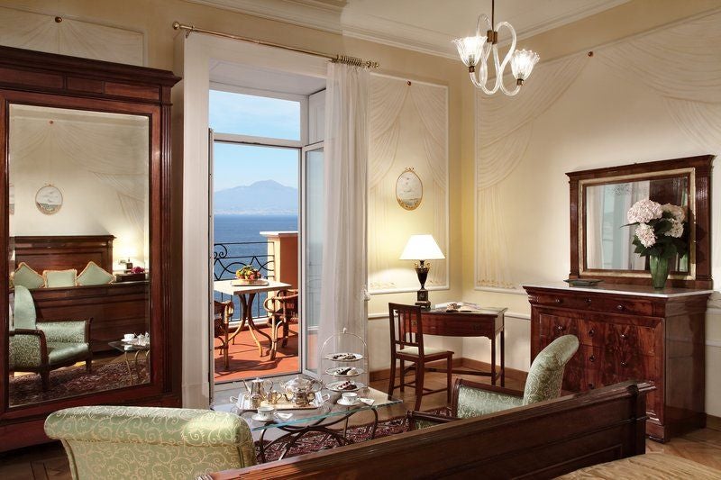 Elegant hotel balcony overlooking the Bay of Naples, with ornate wrought-iron railings, potted plants, and two cushioned chairs for sea views