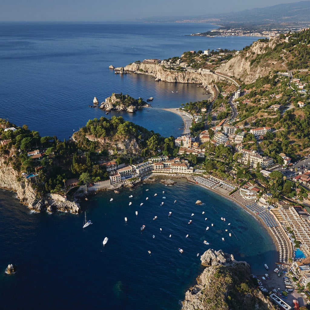 Elegant beachfront hotel with private cabanas overlooking turquoise Mediterranean waters, nestled against Taormina's rocky coastline.