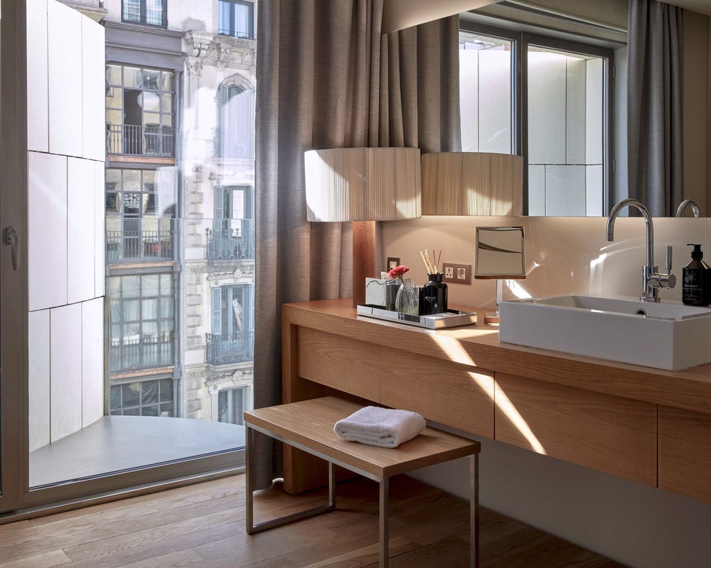 Elegant modern hotel room with minimalist design, warm wood tones, sleek furniture, and large window overlooking urban Spanish cityscape at sunset