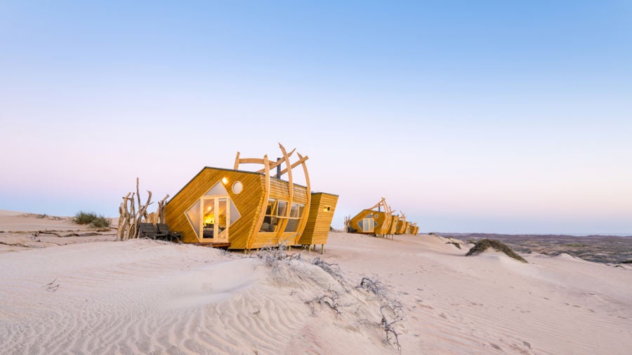 Modern angular cabin perched on sand dunes overlooking Namibia's Skeleton Coast, designed to evoke a weathered shipwreck at sunset