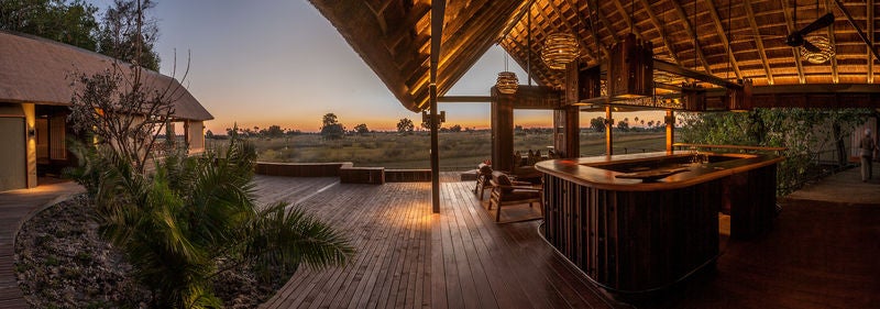 Elevated wooden walkway leads to luxurious safari tents nestled among acacia trees, overlooking a scenic floodplain at sunset