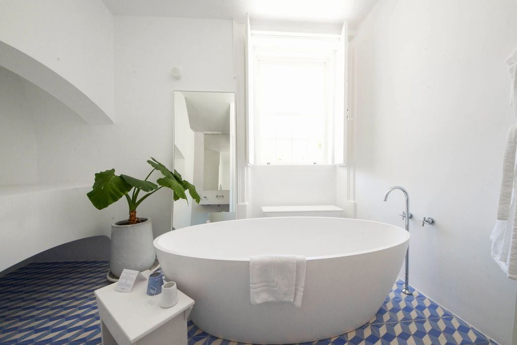 Bright coastal suite with freestanding oval tub, white-draped canopy bed, exposed stone walls and panoramic balcony overlooking Lagos Bay