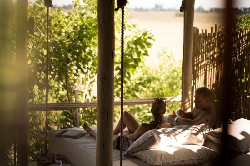 Elevated wooden luxury safari lodge with expansive deck overlooking African savanna grasslands at sunset, canvas tents visible