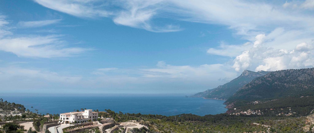 Elegant Spanish luxury hotel nestled in mountains with stone facade, terracotta roof, and manicured gardens overlooking coastline